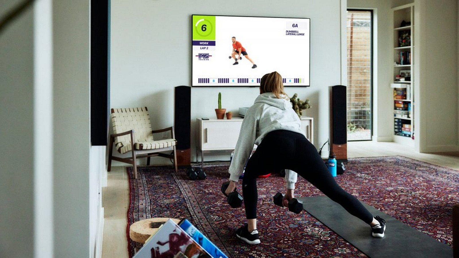 Person performing a lateral lunge exercise while following an F45 Training workout on a Samsung TV in a cozy living room setup, featuring a large rug, chair, and various fitness equipment.